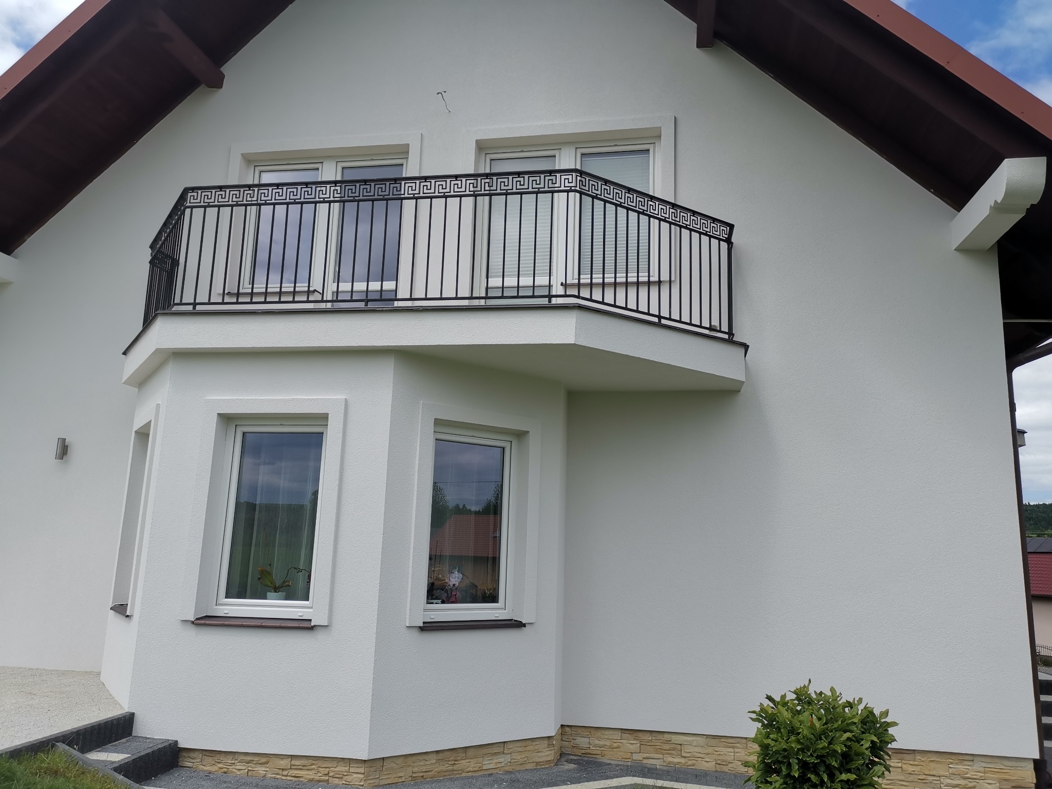 Biały dom z nową elewacją, balkonem z czarną, ozdobną balustradą w stylu greckim i oknami z białymi ramami.