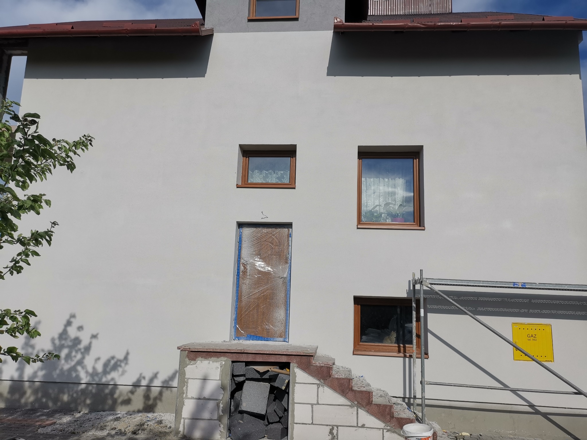 Dwukondygnacyjny dom po szpachlowaniu elewacji, z oknami w brązowych ramach, zabezpieczonymi drzwiami wejściowymi, widoczne rusztowanie i schody z cegły klinkierowej w trakcie budowy.