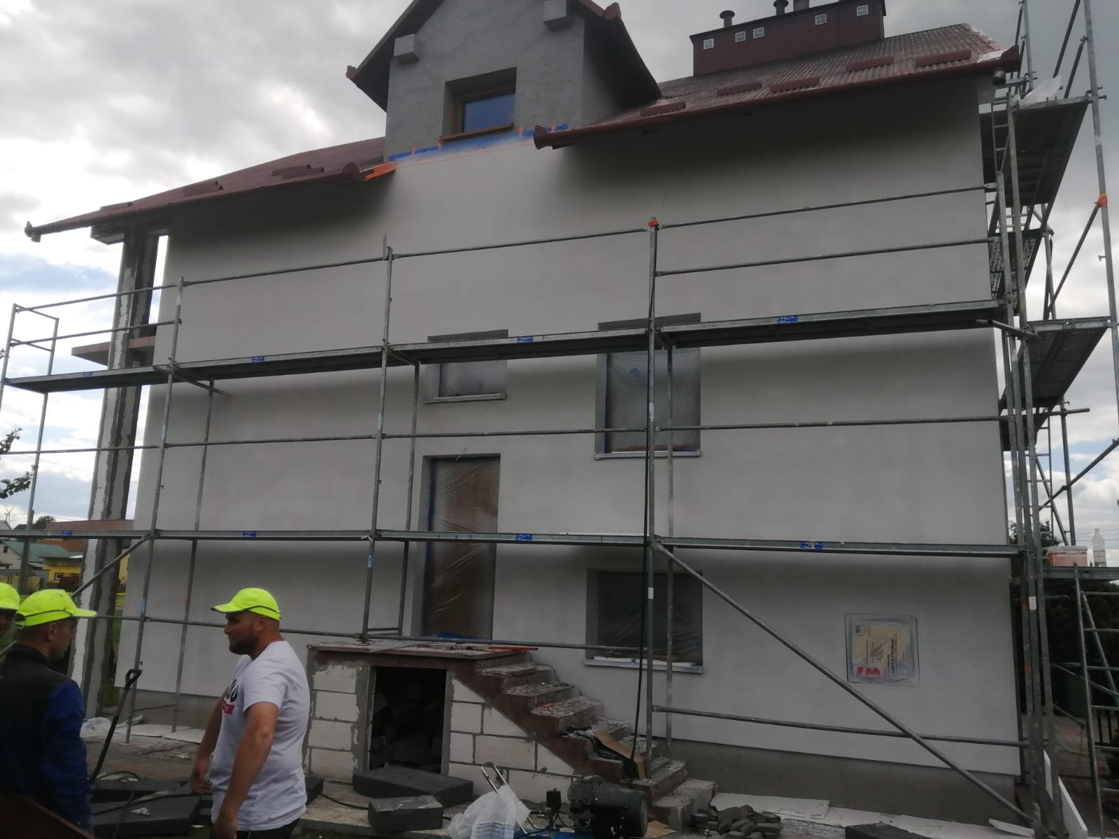 Dwupiętrowy dom w trakcie ocieplania, z ustawionymi rusztowaniami, widoczne zakryte okna folią, pracownicy w neonowych czapkach, szare niebo.