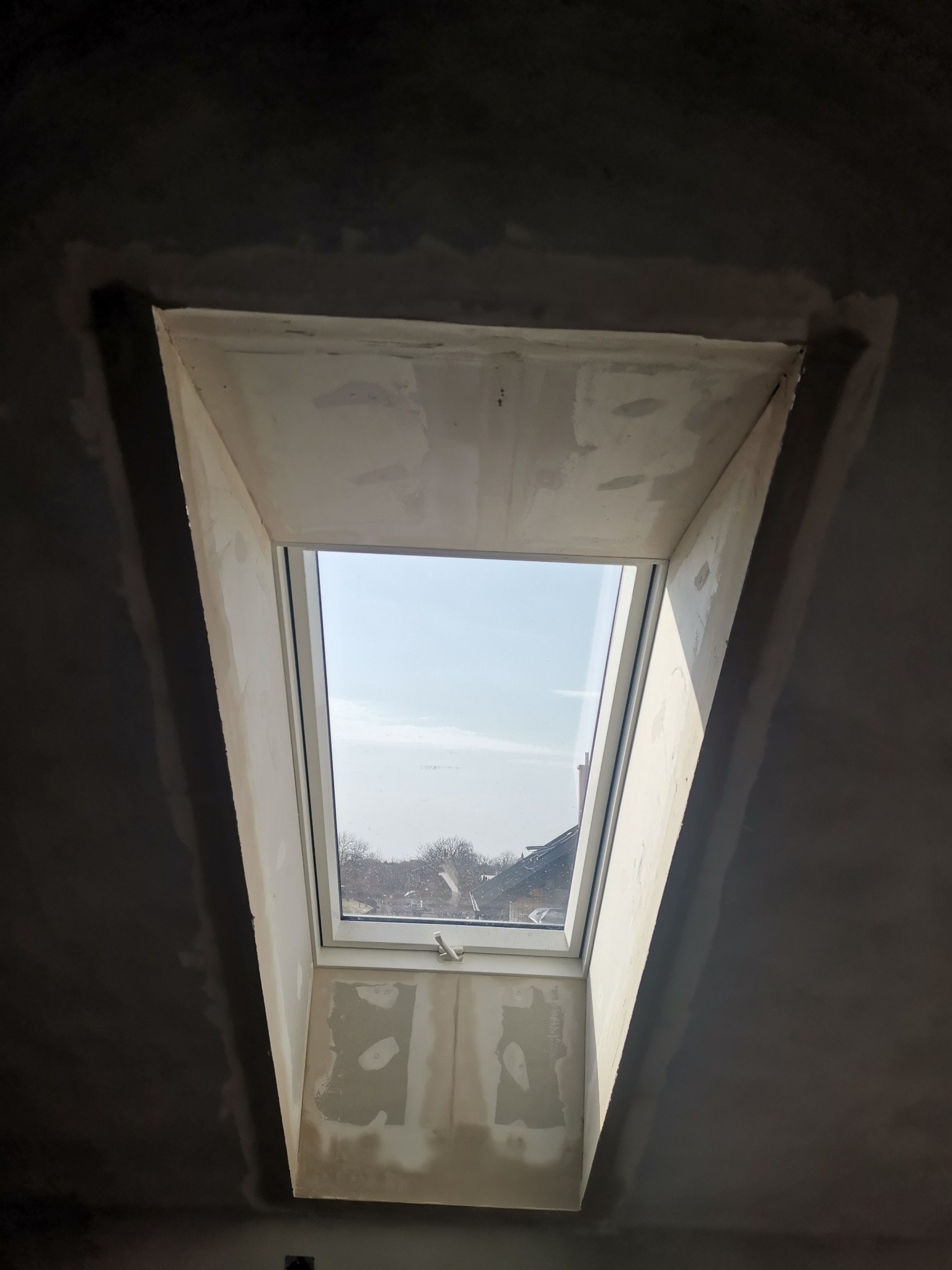 Montaż okna dachowego w surowym wnętrzu, widoczne ślady szpachlowania wokół ramy okiennej i fragment nieotynkowanego sufitu.