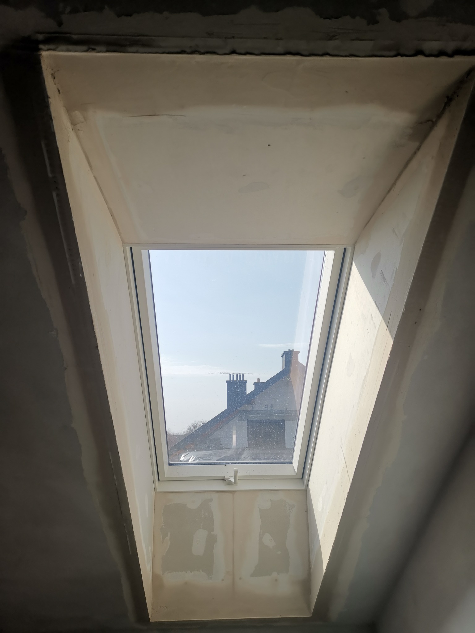 Wnętrze poddasza z oknem dachowym w trakcie szpachlowania, widoczne niedoskonałości powierzchni i częściowo wyschnięta masa szpachlowa, przez okno widać fragment dachu sąsiedniego budynku.