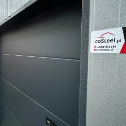 Nowoczesna, grafitowa brama garażowa segmentowa z poziomymi przetłoczeniami, z naklejoną wizytówką firmy coSteel.pl na metalowej ościeżnicy w deszczowy dzień.