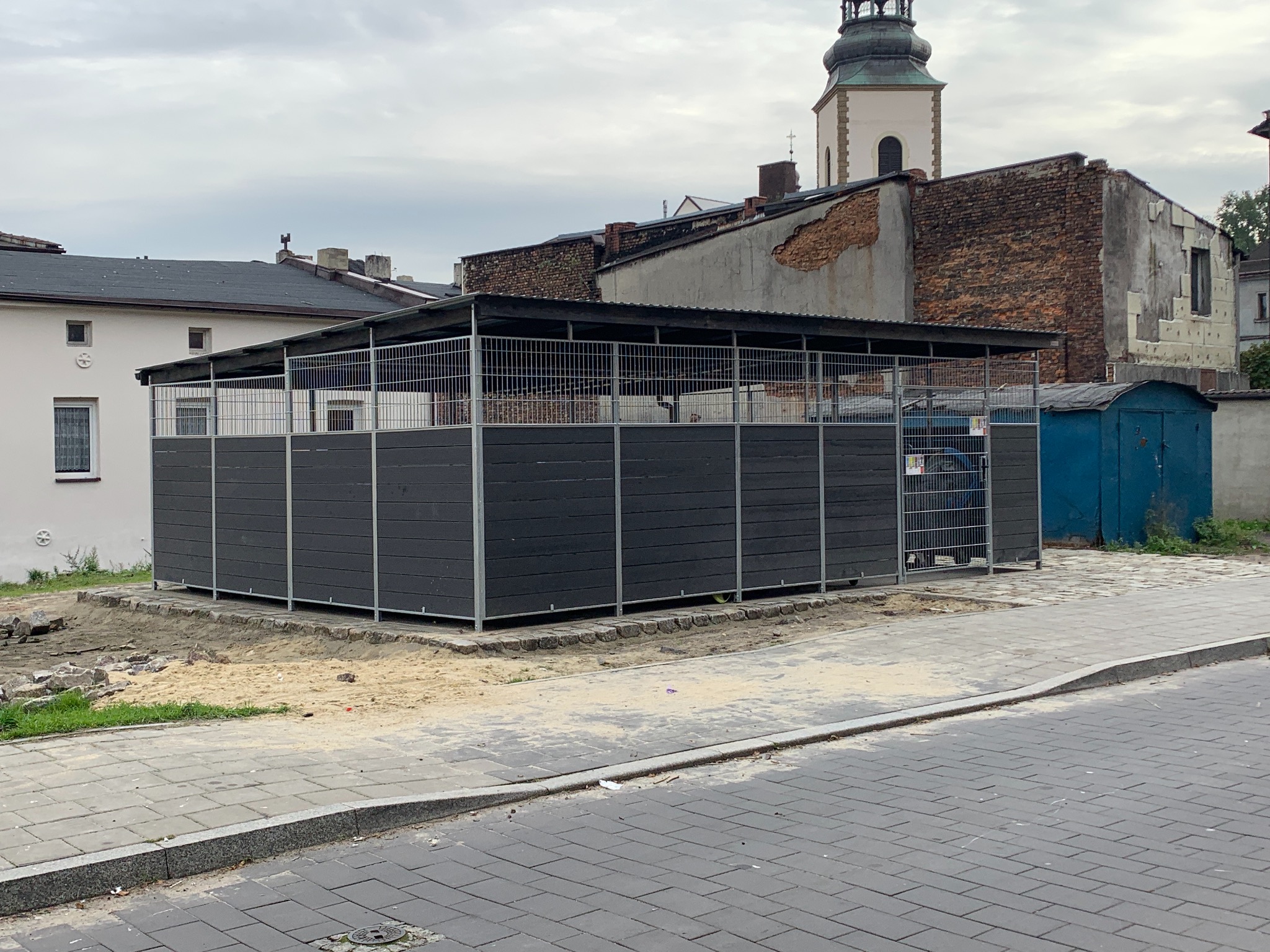 Zewnętrzna konstrukcja z metalową kratą i ciemnymi panelami, prawdopodobnie wiata śmietnikowa, na tle miejskiego krajobrazu z kościelną wieżą.