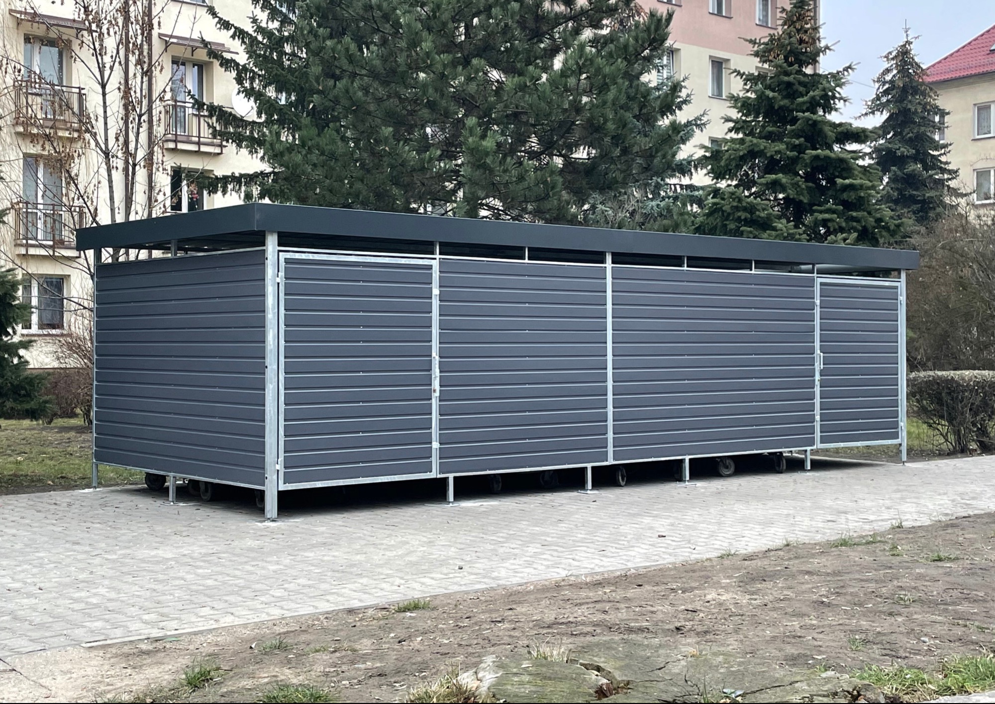 Szara, modułowa wiata śmietnikowa z poziomych paneli, na kółkach, na tle bloku mieszkalnego i drzew.