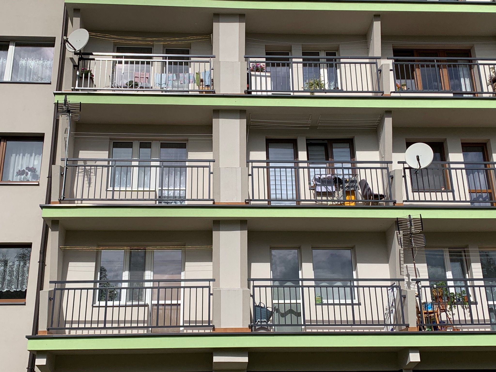 Widok z dołu na fasadę budynku wielorodzinnego z balkonami zabezpieczonymi metalowymi barierkami; na niektórych balkonach widoczne suszarki na pranie, kwiaty i anteny satelitarne.