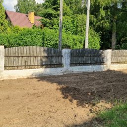 Henrix Dorota Krulikowska - Przygotowany teren pod trawnik, wyrównana ziemia z widocznym ogrodzeniem z drewnianymi panelami i betonowymi słupkami, w tle zieleń drzew i krzewów oraz dom z brązowym dachem.