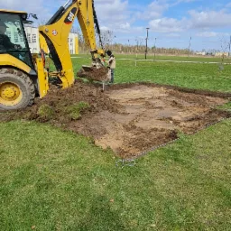 Żółta koparko-ładowarka CAT podczas prac ziemnych, wykop pod przyszłą konstrukcję, widoczne wytyczone granice terenu sznurkiem, trawa i błękitne niebo w tle.