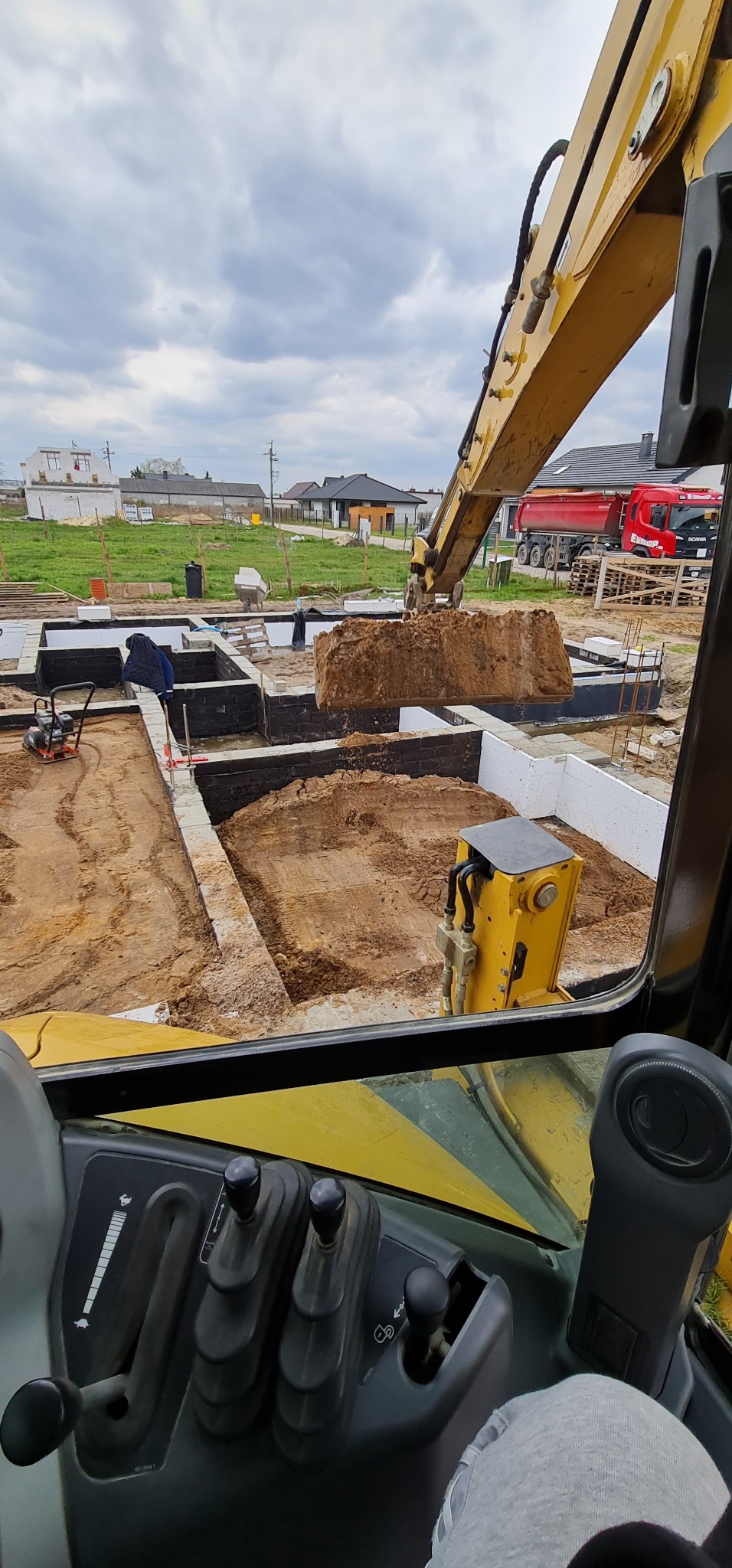 Widok z kabiny koparki podczas zasypywania fundamentów piaskiem; w tle widoczny budynek w trakcie budowy oraz czerwona ciężarówka.