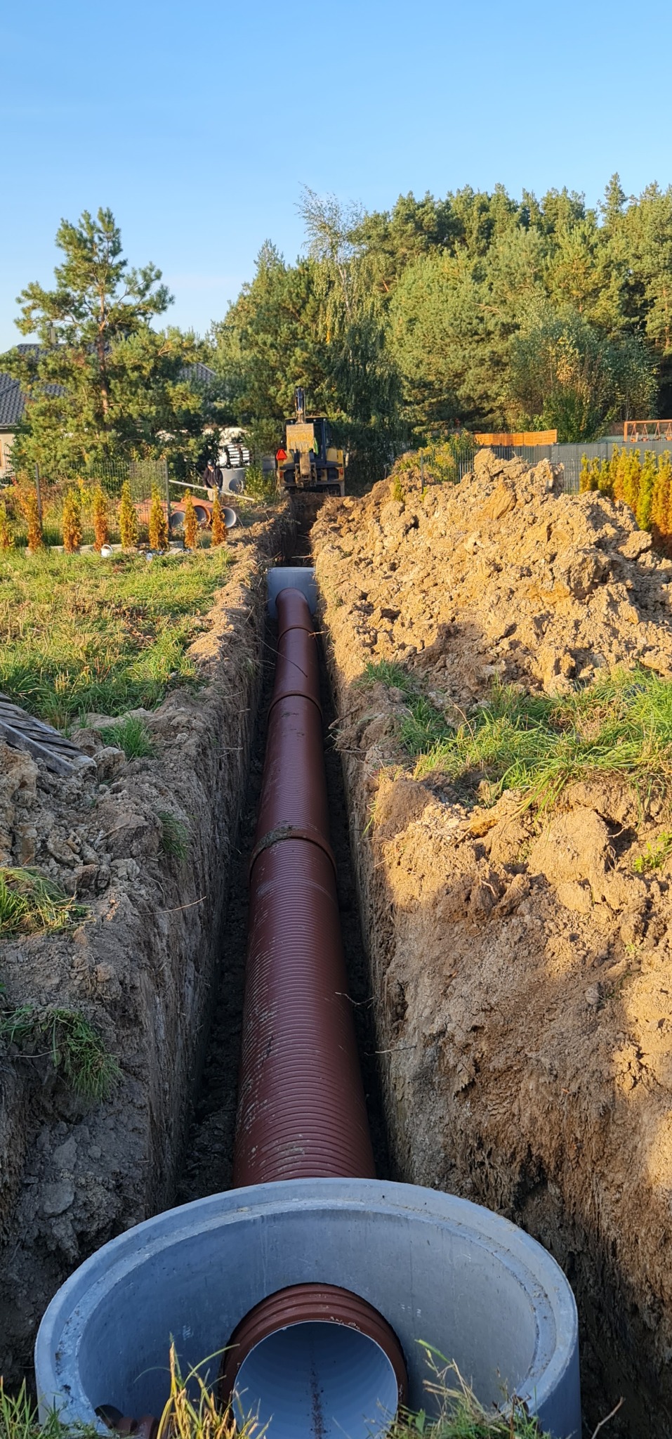 Brązowa rura kanalizacyjna ułożona w wykopie, widoczny betonowy element studni, w tle koparka i zieleń.