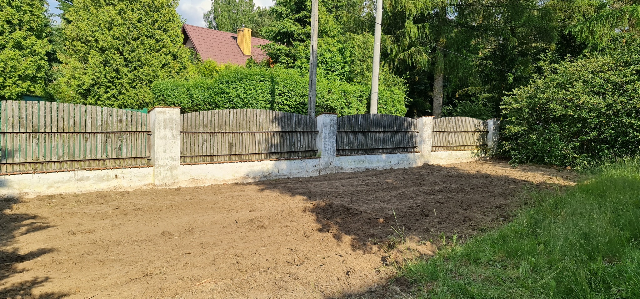 Przygotowany teren pod trawnik, wyrównana ziemia z widocznym ogrodzeniem z drewnianymi panelami i betonowymi słupkami, w tle zieleń drzew i krzewów oraz dom z brązowym dachem.