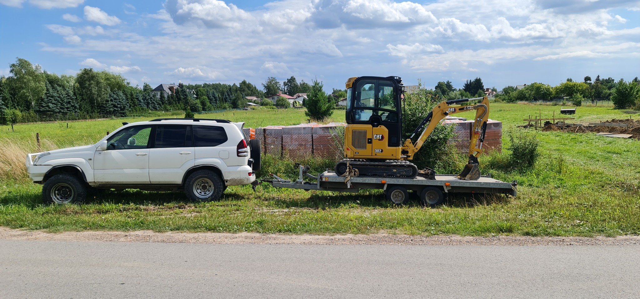Biały samochód terenowy ciągnie przyczepę z małą żółtą koparką CAT 302 CR. W tle widoczny teren budowy z przygotowanymi fundamentami oraz palety z cegłami. Krajobraz wiejski z domami i drzewami...