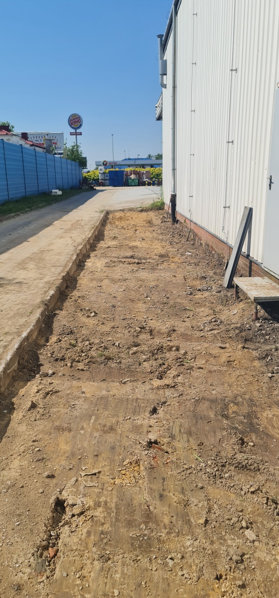 Przygotowany teren pod drogę lub chodnik, widoczne ślady po maszynach, obok budynek przemysłowy i reklama Burger King w tle.