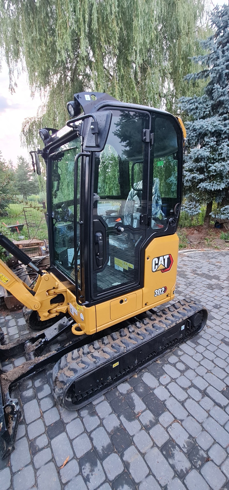 Żółto-czarna mini koparka CAT 302 zaparkowana na szarej kostce brukowej w Nadarzynie, z zielonymi drzewami w tle.