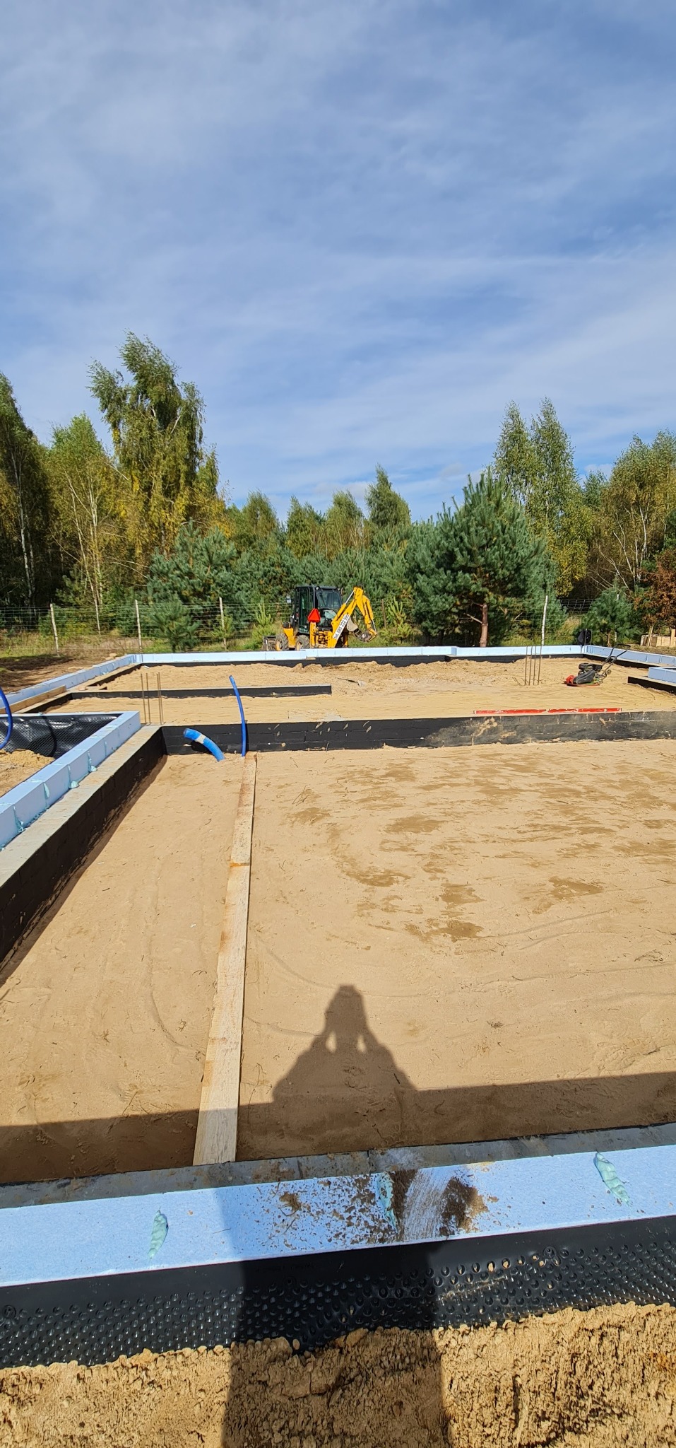 Wykop pod fundamenty z widoczną izolacją pionową, szalunkiem ze styropianu i pracującą koparką w tle, otoczony drzewami.