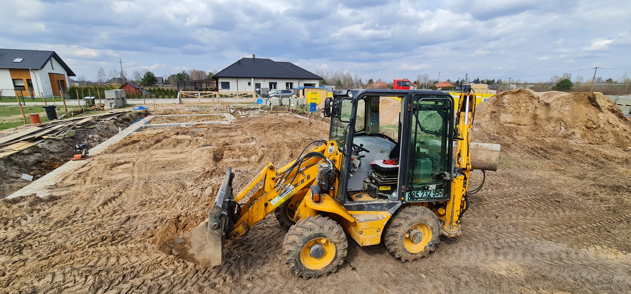 Żółta mini ładowarka JCB na placu budowy, widoczne wykopane fundamenty domu jednorodzinnego i pryzma piasku, w tle zabudowa mieszkalna i pochmurne niebo.
