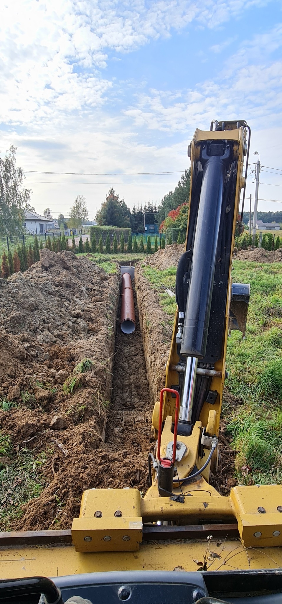 Żółta koparka z przodu, w tle wykopany rów z ułożonymi rurami kanalizacyjnymi, widok na ogród z tujami i zabudowania w oddali, pochmurne niebo.