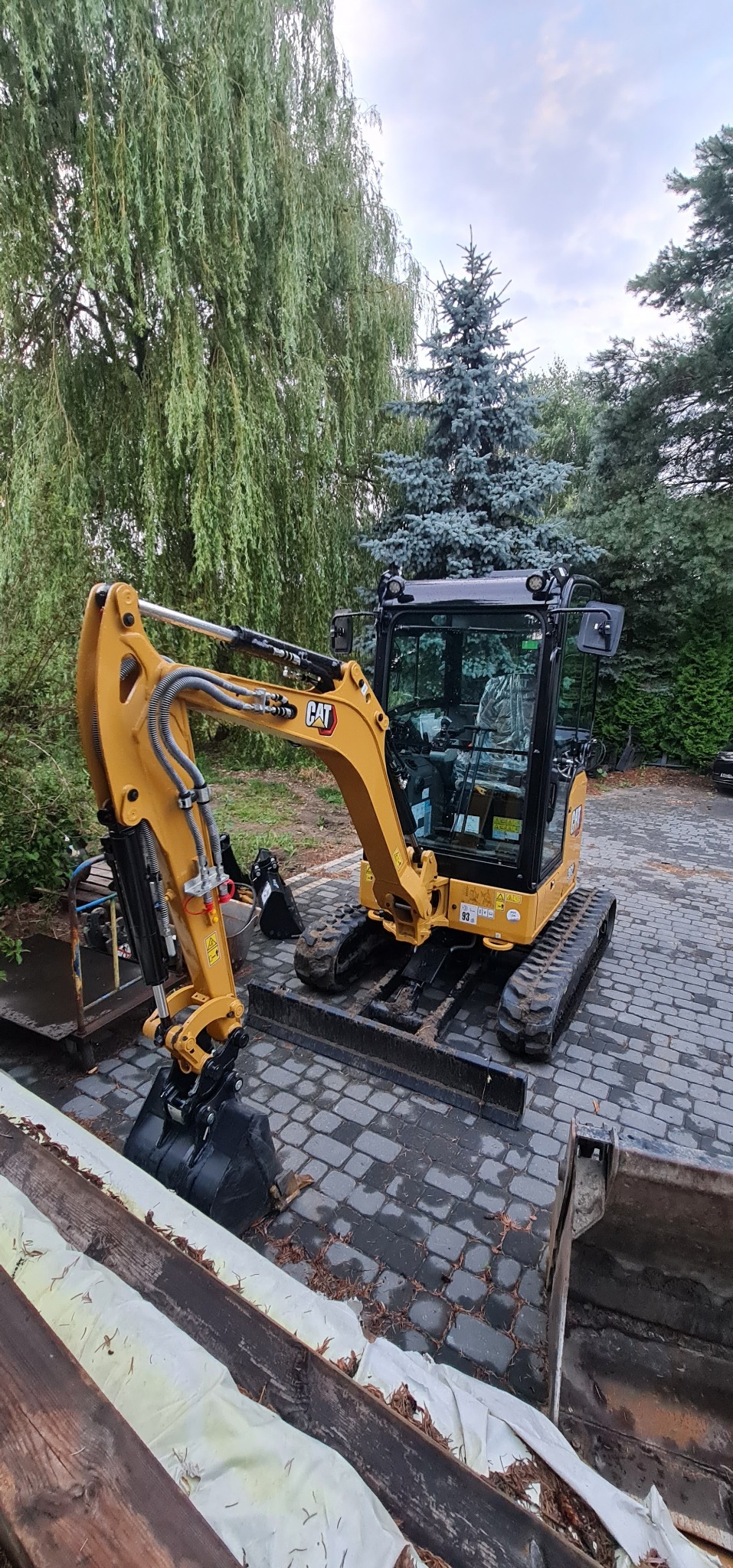 Żółta mini koparka CAT na kostce brukowej, z widocznymi przewodami hydraulicznymi i łyżką, w tle drzewa iglaste i liściaste.
