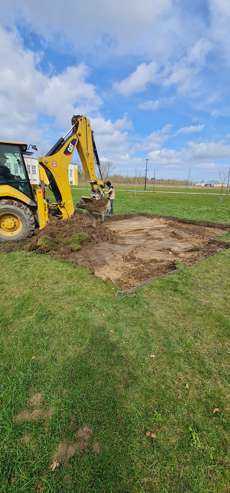 Żółta koparko-ładowarka CAT podczas prac ziemnych, wykop pod przyszłą konstrukcję, widoczne wytyczone granice terenu sznurkiem, trawa i błękitne niebo w tle.