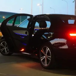 Czarny samochód osobowy marki Mercedes z otwartymi drzwiami, widoczne podświetlenie wnętrza i tylne światła, na tle budynku z zielonym oświetleniem.