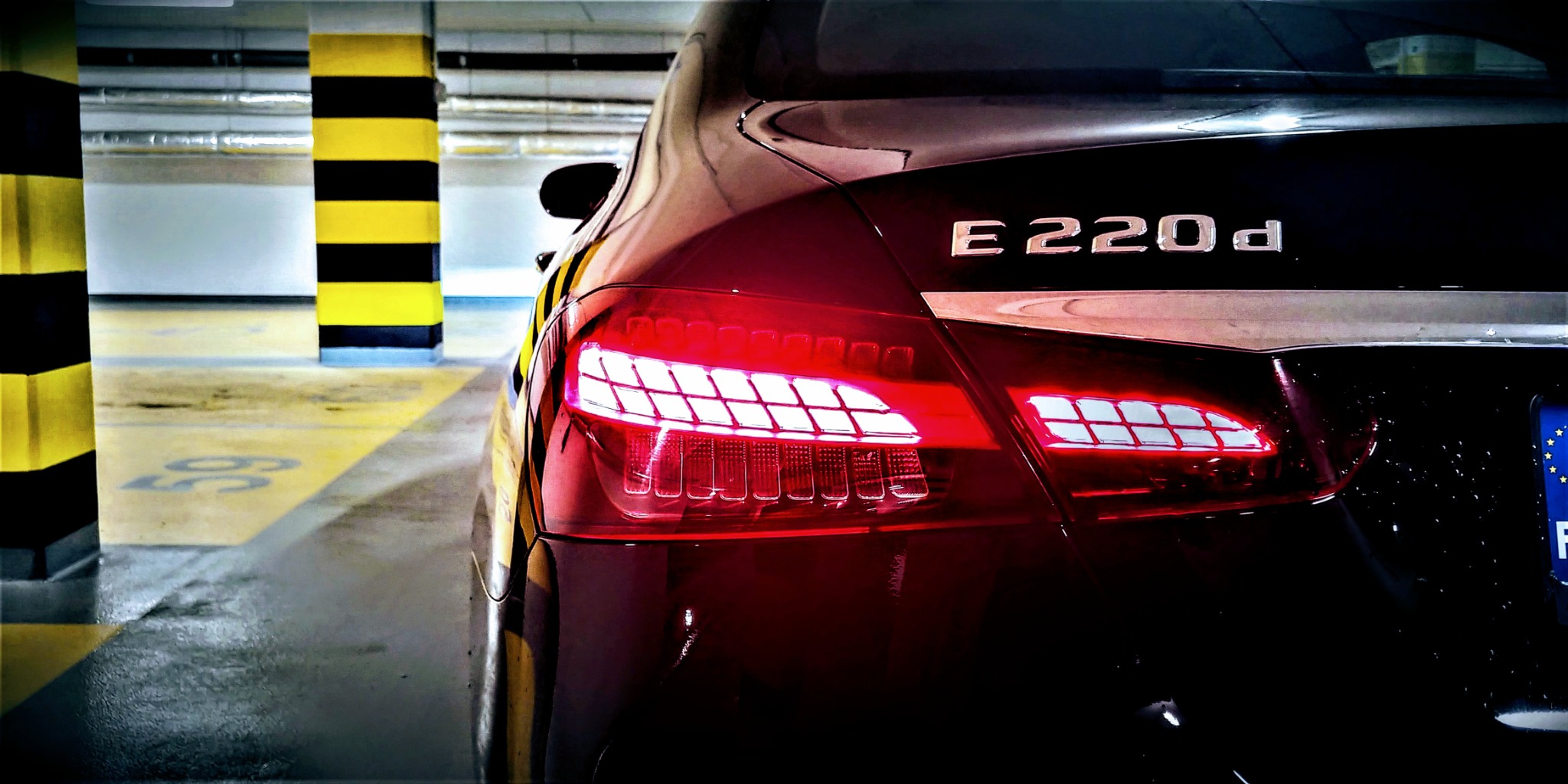 Tył czarnego samochodu Mercedes-Benz E220d z włączonymi światłami, sfotografowany w słabo oświetlonym garażu podziemnym z żółto-czarnymi słupami.