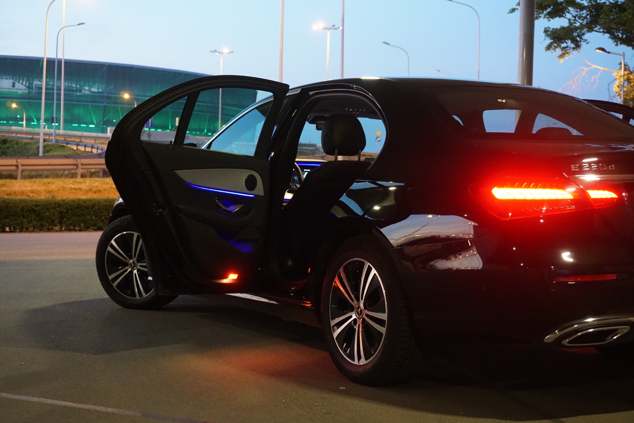 Czarny samochód osobowy marki Mercedes z otwartymi drzwiami, widoczne podświetlenie wnętrza i tylne światła, na tle budynku z zielonym oświetleniem.