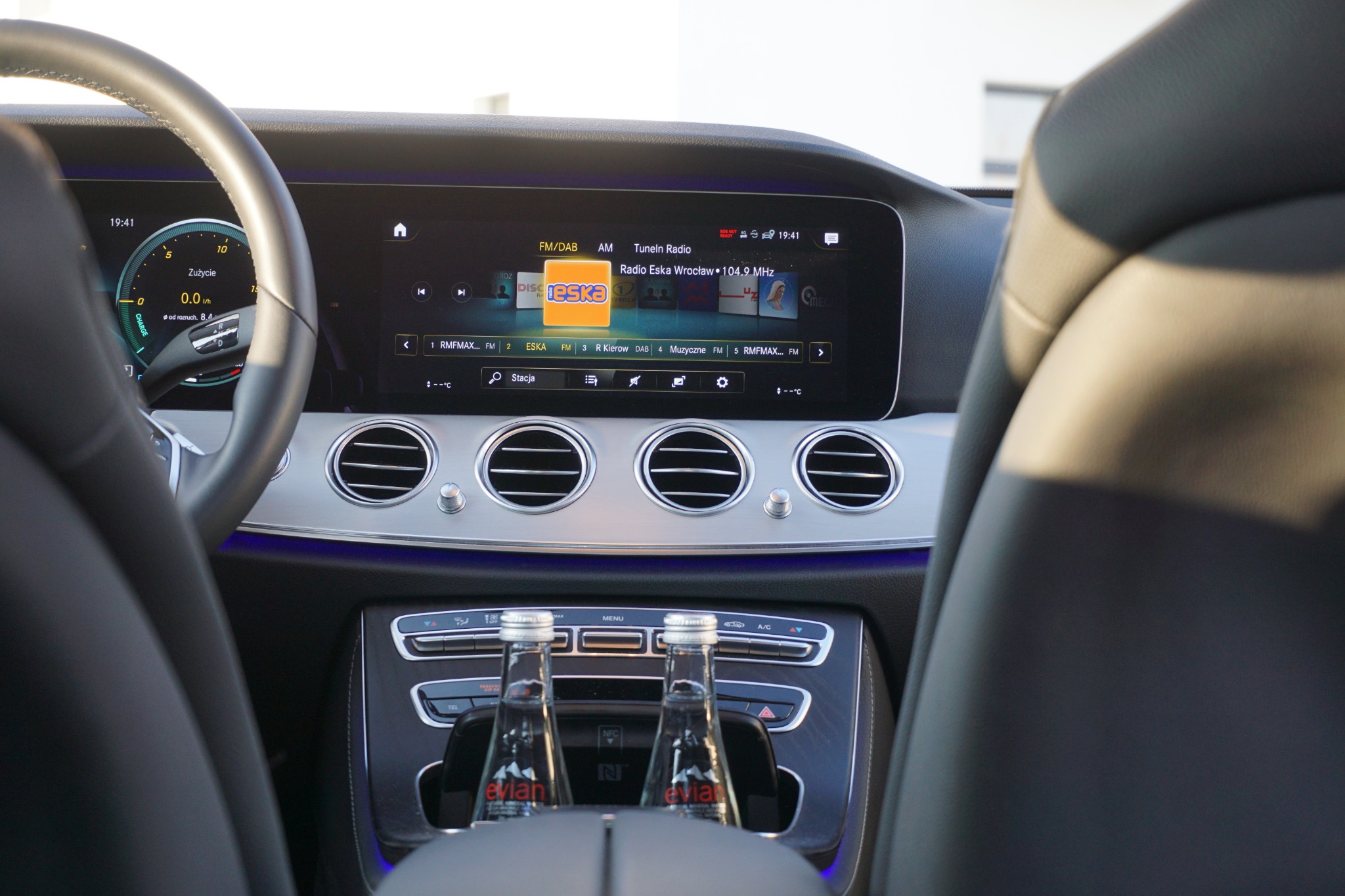 Wnętrze samochodu z widocznym radiem Eska Wrocław na wyświetlaczu, srebrnymi nawiewami i butelkami wody Evian w uchwycie.