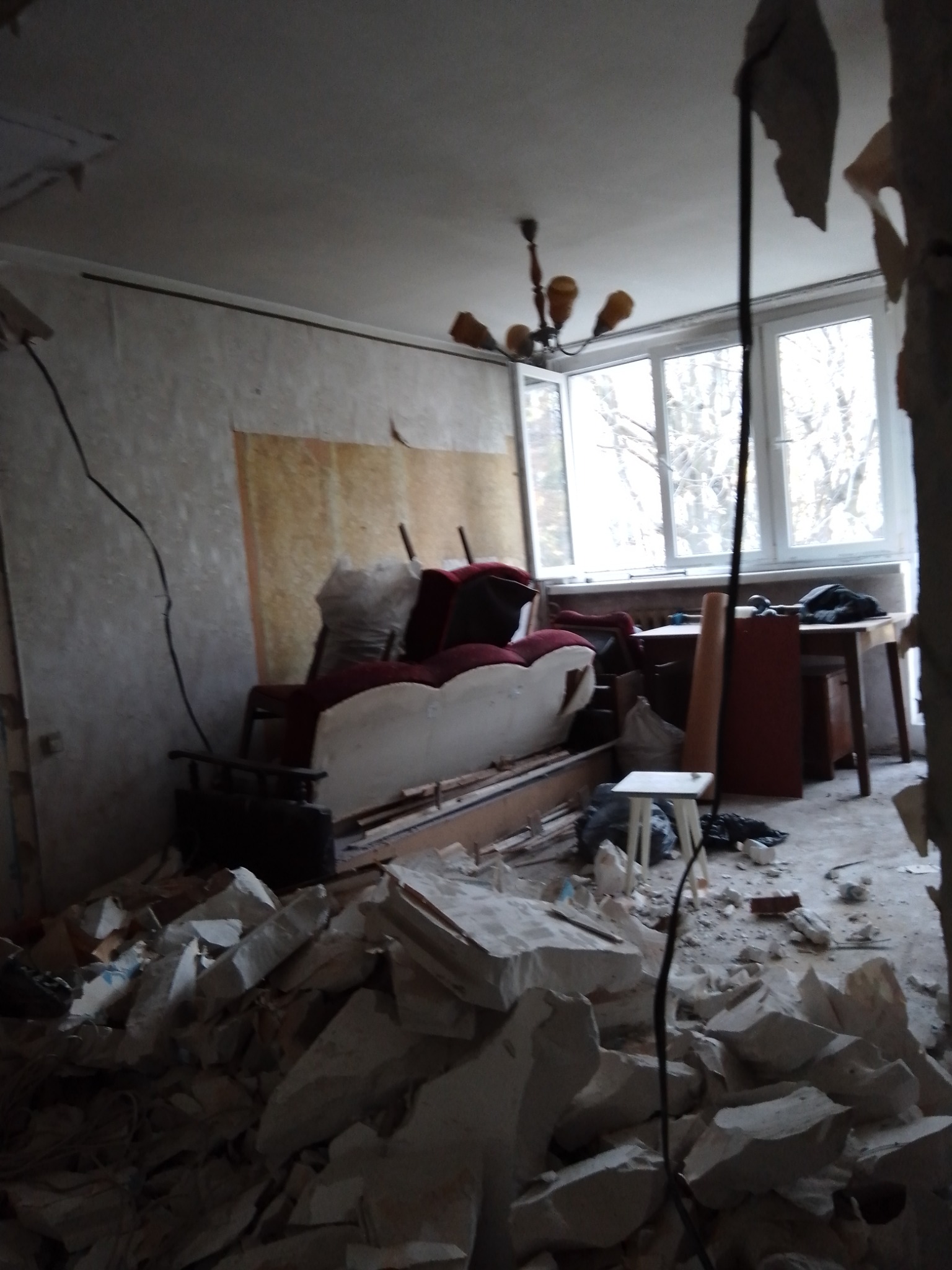 Wnętrze pokoju w trakcie rozbiórki, z gruzem na podłodze, odsłoniętą ścianą i widocznym fragmentem sofy oraz mebli w tle, oświetlone światłem dziennym wpadającym przez okno.