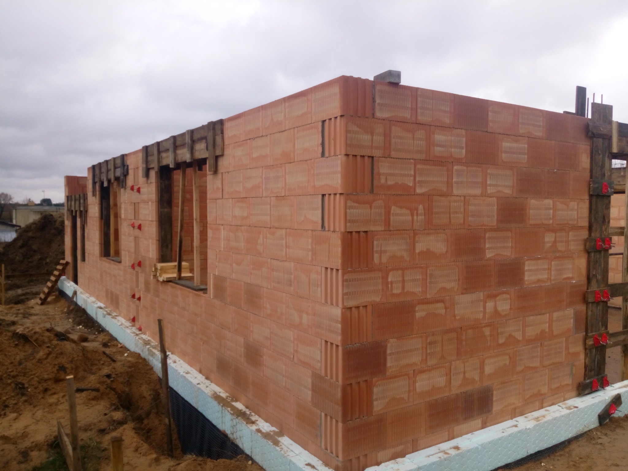 Wznoszenie ścian parteru z pustaków ceramicznych, widoczne deskowanie otworów okiennych i izolacja fundamentów styropianem.