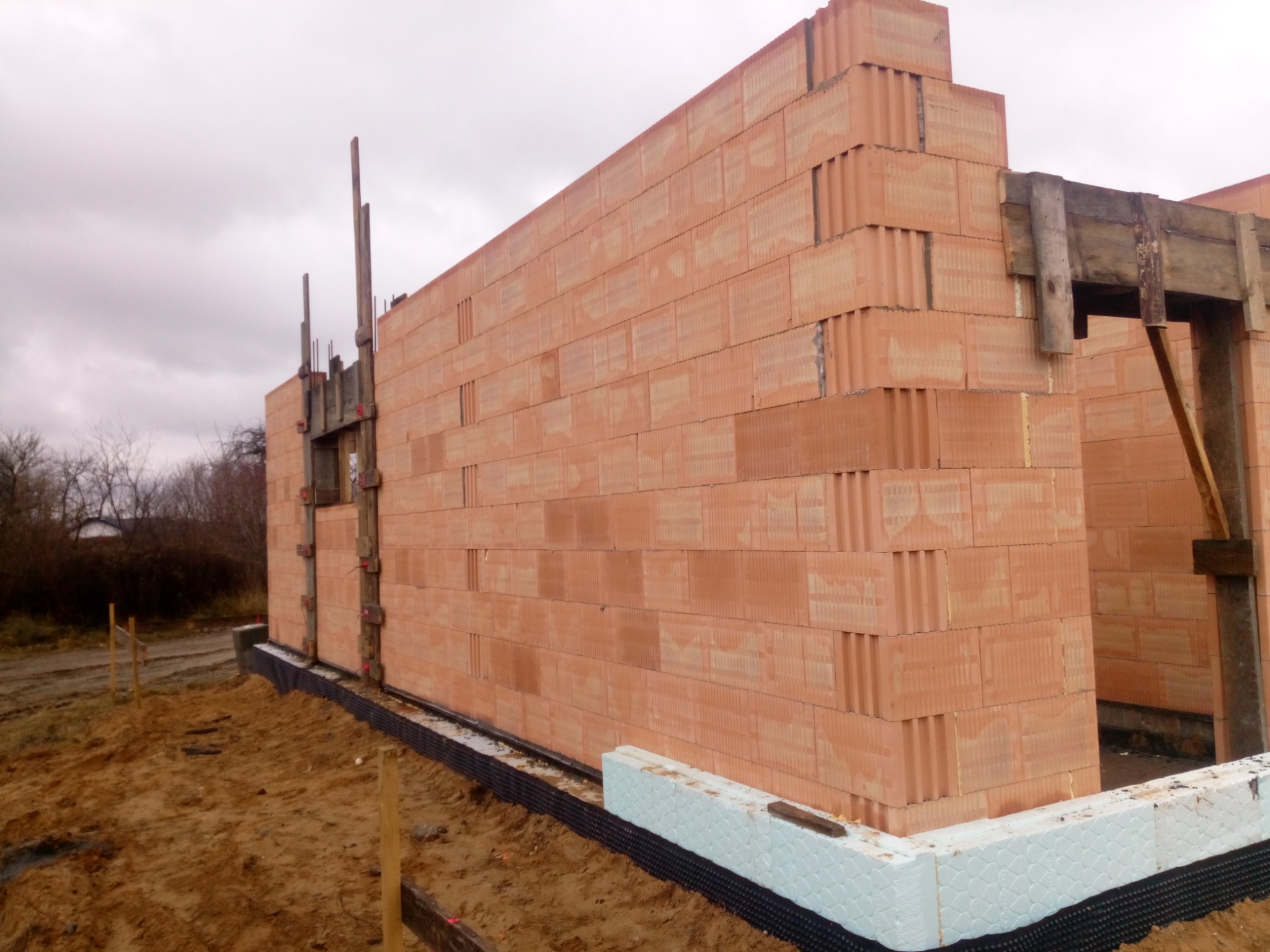 Wznoszone ściany domu murowanego z czerwonej ceramiki poryzowanej, widoczna izolacja fundamentów styropianem i folią kubełkową, drewniane stemple podtrzymujące nadproże.