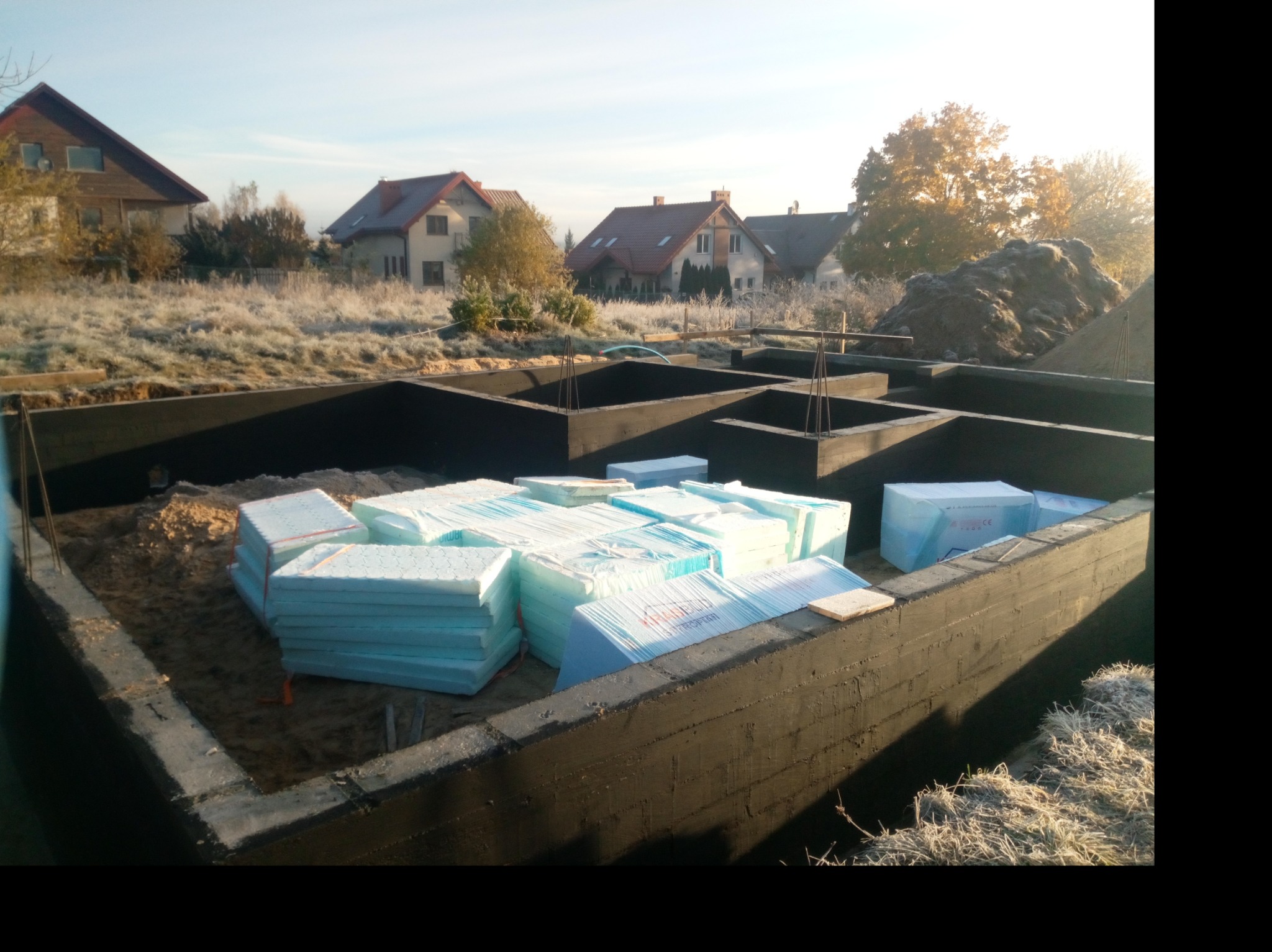 Fundamenty domu jednorodzinnego z hydroizolacją, przygotowane do ocieplenia styropianem, z widocznymi domami w tle.