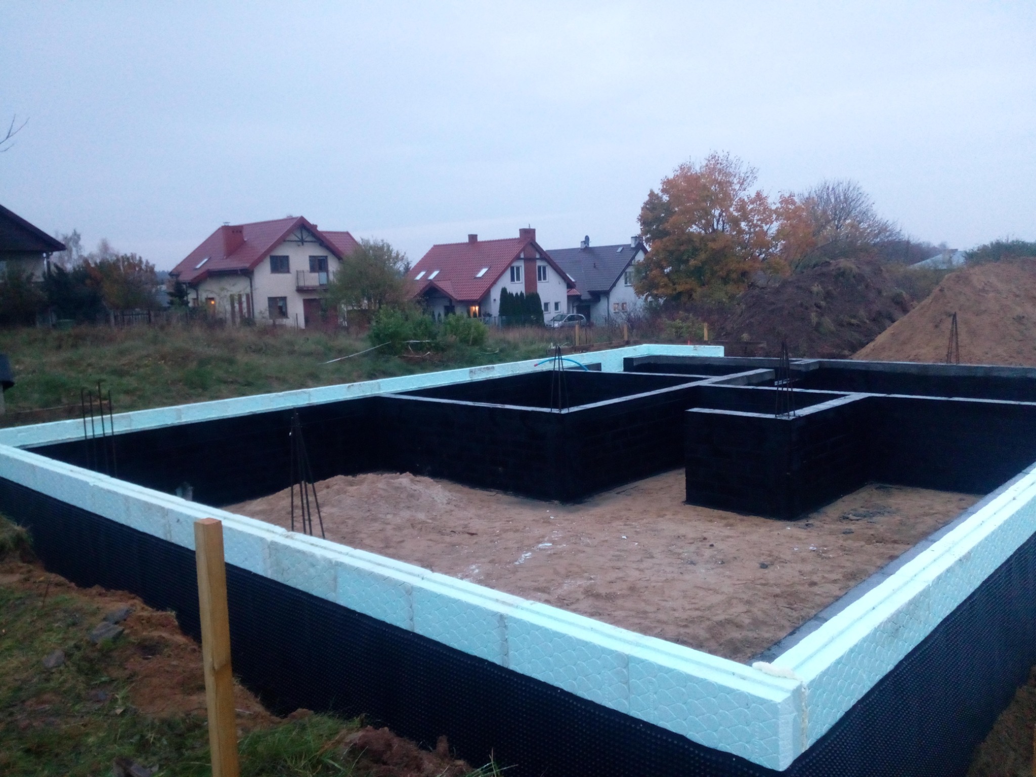 Przygotowany fundament z izolacją termiczną i przeciwwilgociową, widoczne wystające pręty zbrojeniowe, w tle zabudowa jednorodzinna w Hryniewiczach.