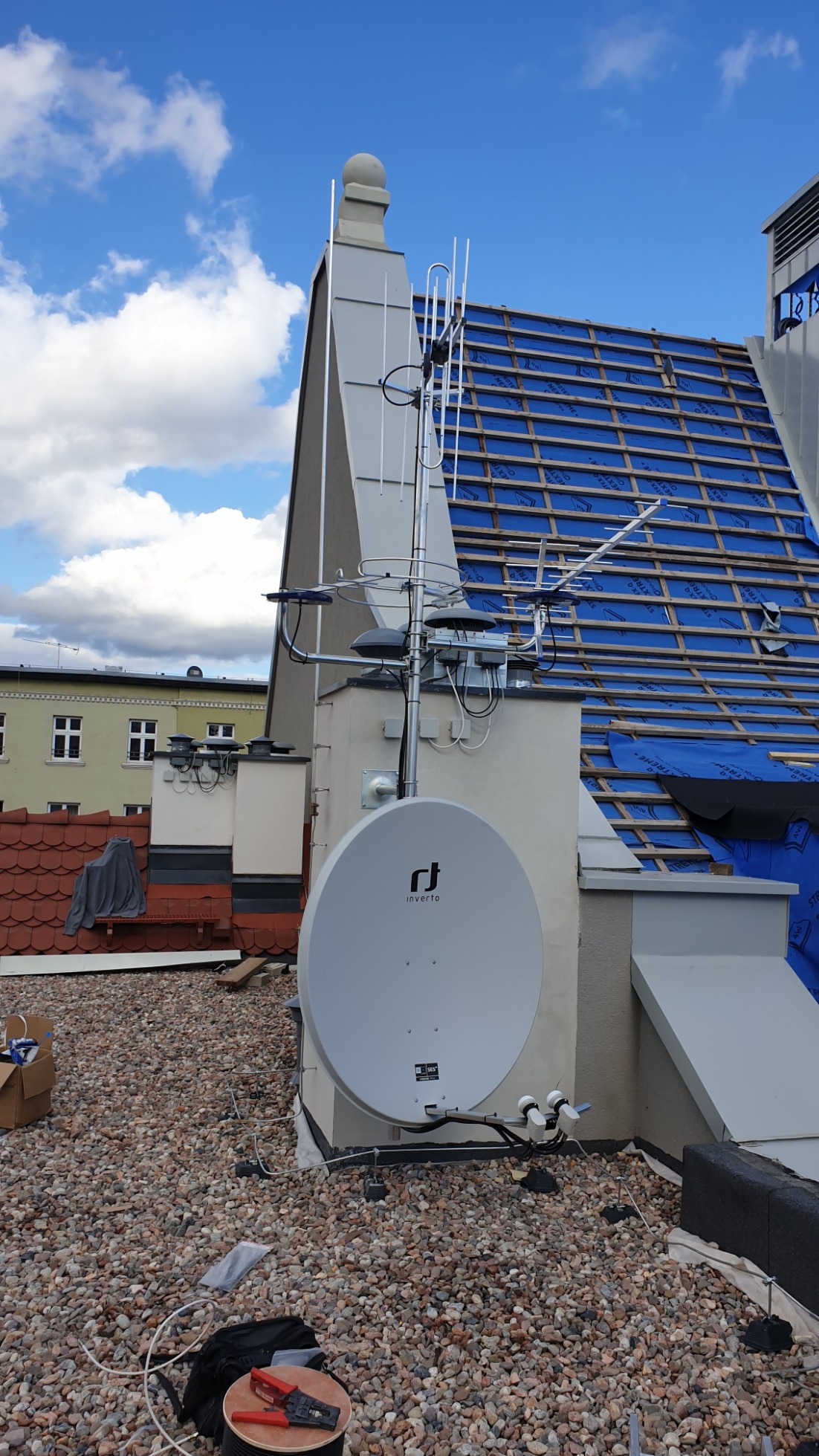Antena satelitarna i antena naziemna zamontowane na dachu budynku z widocznymi narzędziami i okablowaniem. Dach pokryty drobnymi kamieniami, w tle budynek w trakcie remontu dachu.