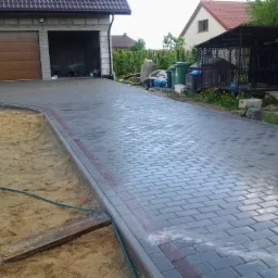 Świeżo ułożona, szara kostka brukowa na podjeździe do garażu, z bordiurą z czerwonej kostki, obok widoczny fragment wykopu i zielony wąż ogrodowy.