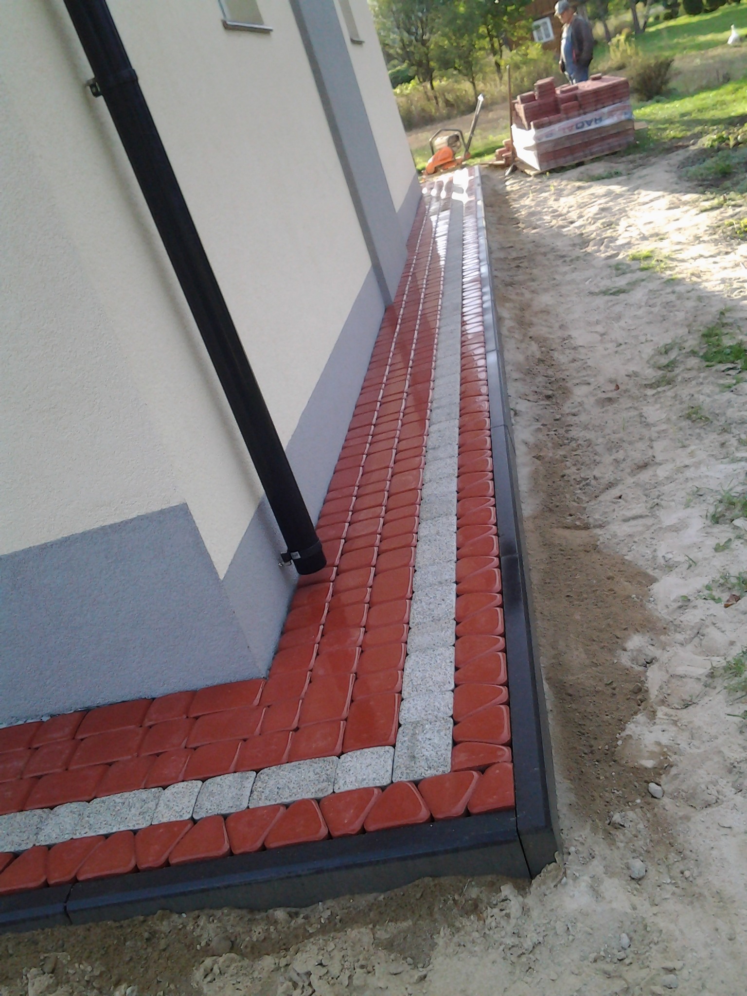 Czerwono-szara kostka ułożona wzdłuż ściany budynku z czarną rynną, widoczny fragment trawnika i ułożone kostki na palecie w tle.