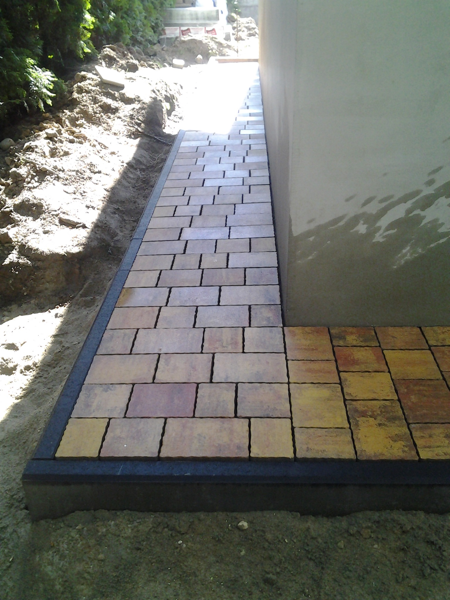 Ścieżka z kostki brukowej w trakcie układania, widoczne krawężniki, ziemia po bokach, fragment budynku z szarą ścianą w tle.