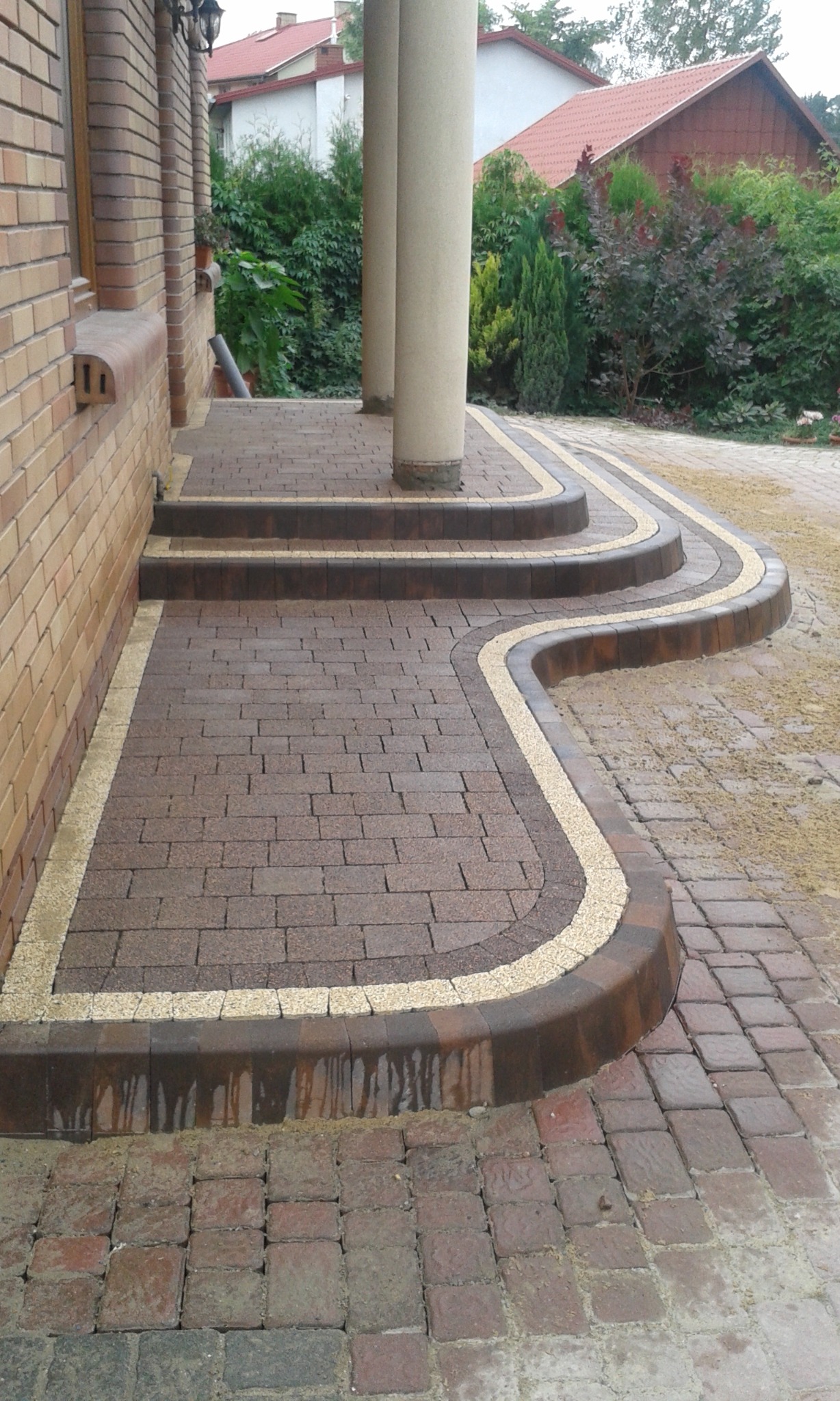 Schody wejściowe z brązowej kostki brukowej z obrzeżem z jasnego grysu, prowadzące do kolumnowego wejścia, widoczne ślady wilgoci na kostce.