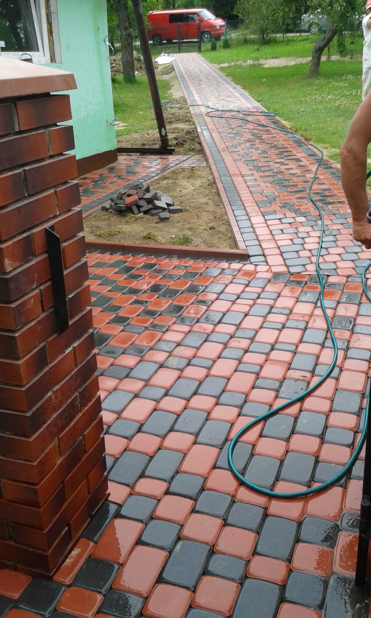 Świeżo ułożona, mokra nawierzchnia z kostki w kolorze czerwono-szarym, ułożonej w romby, widoczny fragment muru z cegły i wąż ogrodowy, w tle czerwony samochód dostawczy.