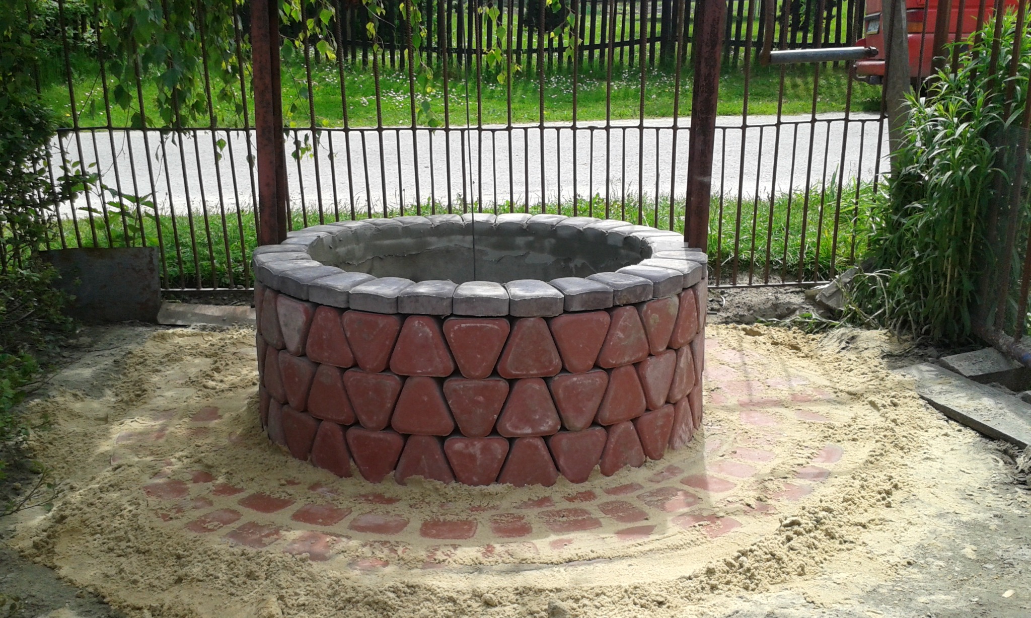 Studnia z czerwonej kostki brukowej o trójkątnym kształcie, otoczona piaskiem, z widoczną metalową bramą w tle.