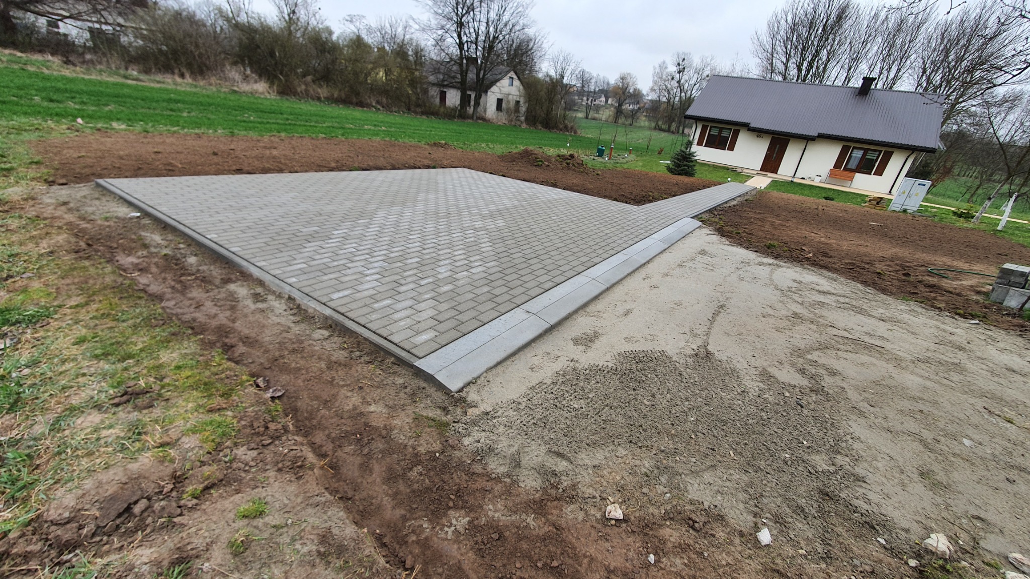 Nowo ułożona, szara kostka brukowa tworząca plac przed domem z widocznymi obrzeżami, przygotowanym podłożem i trawnikiem w tle.