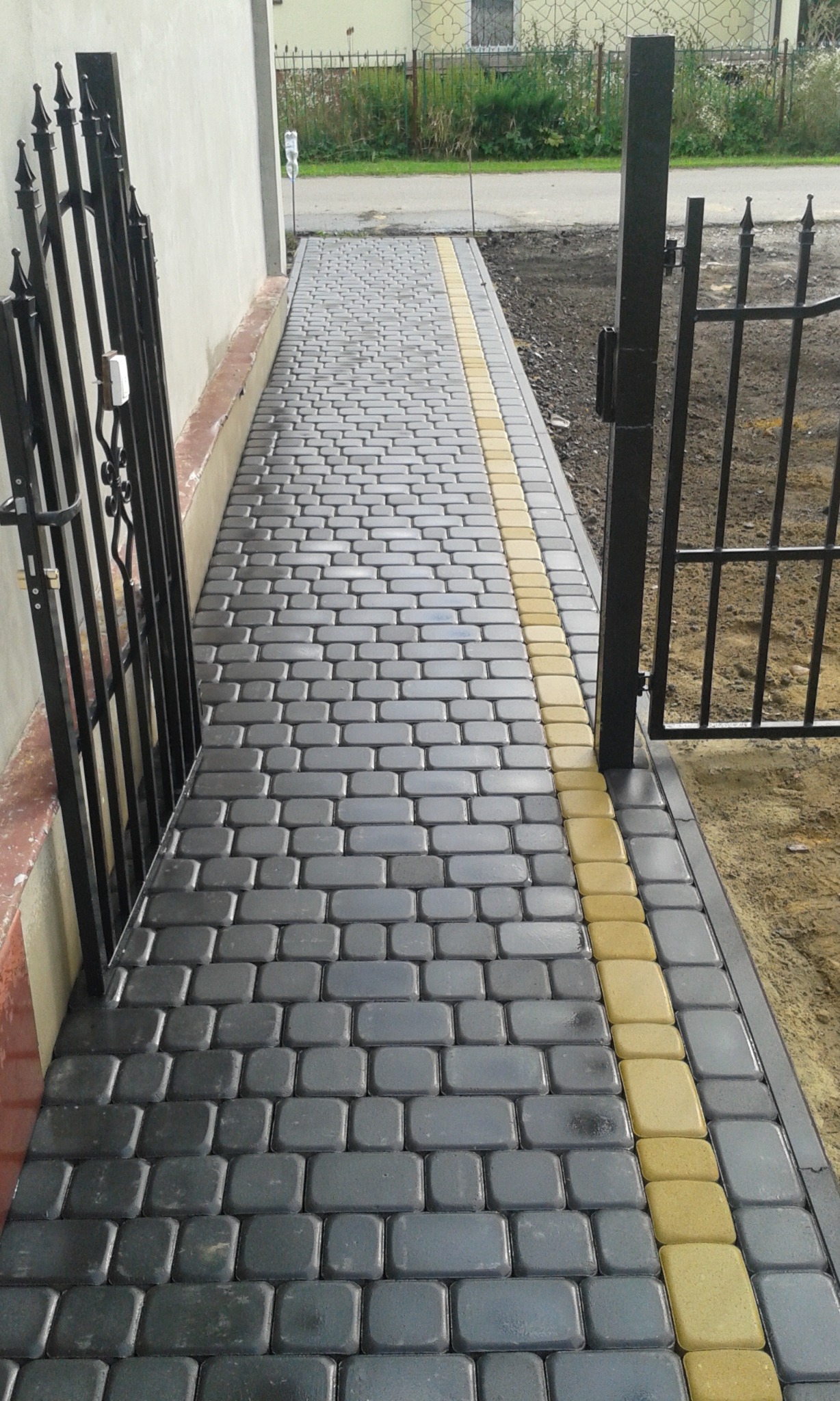 Ułożona ścieżka z szarej kostki brukowej z żółtymi obrzeżami, prowadząca od furtki do budynku.