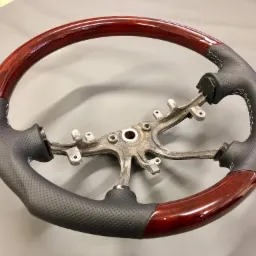Odnowiona kierownica samochodowa: drewniany wieniec z mahoniowym połyskiem, obszyty czarną perforowaną skórą z białymi szwami, metalowy szkielet.
