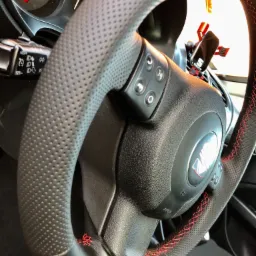 Czarna, perforowana skórzana kierownica samochodowa z czerwonym, ozdobnym szwem krzyżykowym, widoczne przyciski sterowania i logo marki.