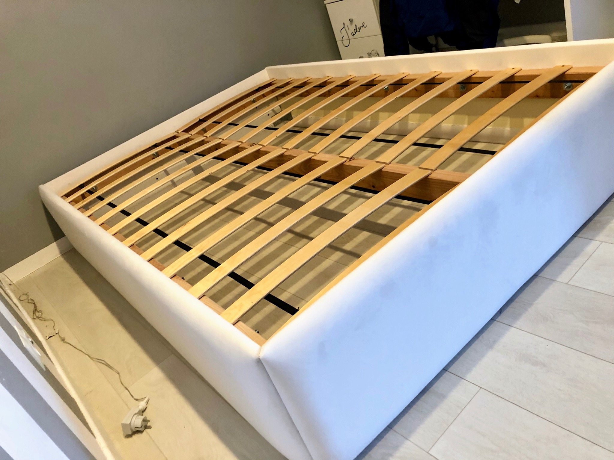 Rama łóżka tapicerowanego białym materiałem, widoczny stelaż z drewnianych listew oraz fragment podłogi z paneli i ściany w tle.