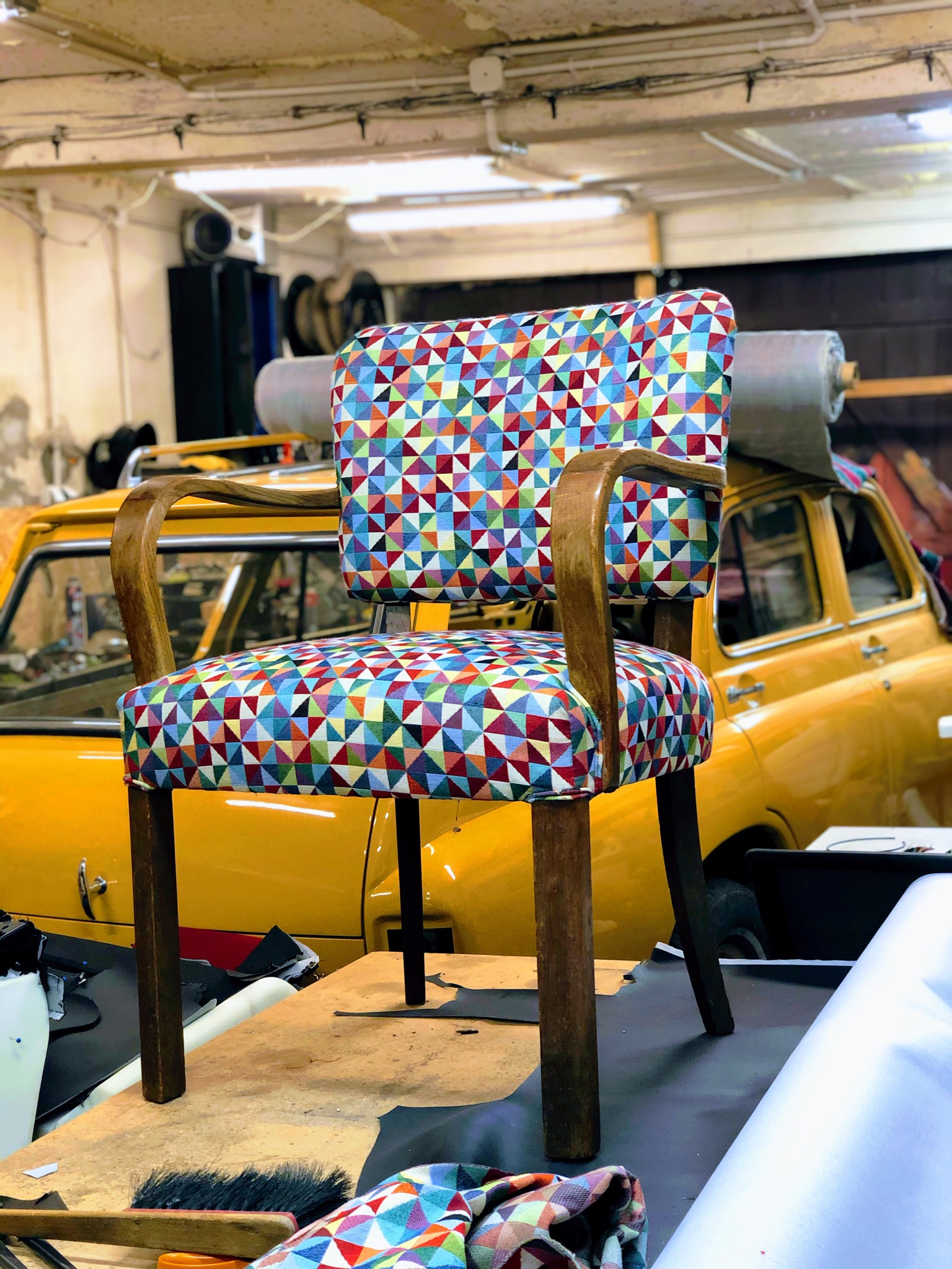 Krzesło z drewnianymi podłokietnikami i siedziskiem obitym tkaniną w kolorowe trójkąty, ustawione na stole w warsztacie, z żółtym samochodem w tle.