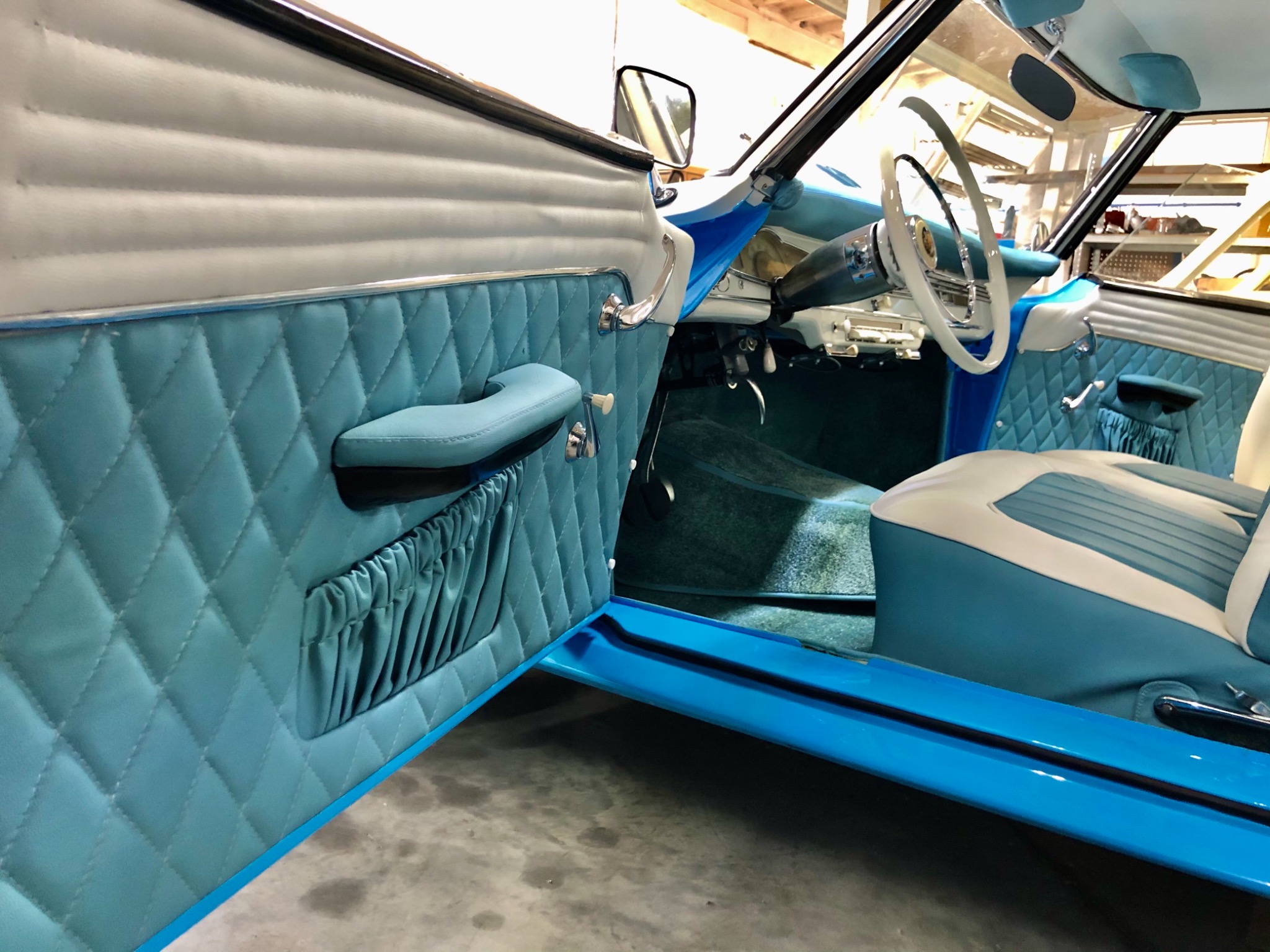 Wnętrze odrestaurowanego samochodu z turkusowo-białą tapicerką w romby na drzwiach i siedzeniach, widoczne detale kokpitu i kierownica.