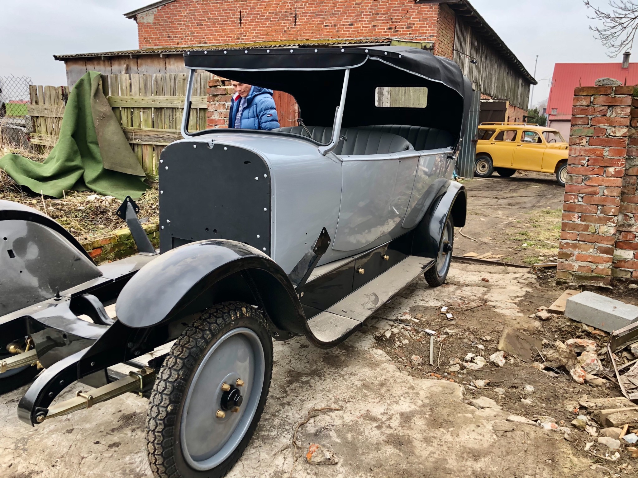 Szary, odrestaurowywany kabriolet z czarnym dachem na zewnątrz, z widoczną ramą i elementami tapicerki, w tle budynki gospodarcze i żółty samochód.