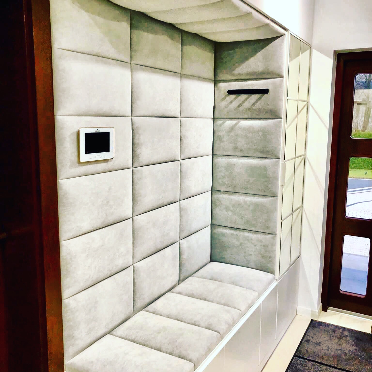 Wnęka wejściowa z miękką, jasnoszarą tapicerką w geometryczne kwadraty, zabudowana siedziskiem i panelem z domofonem, obok drewniane drzwi.