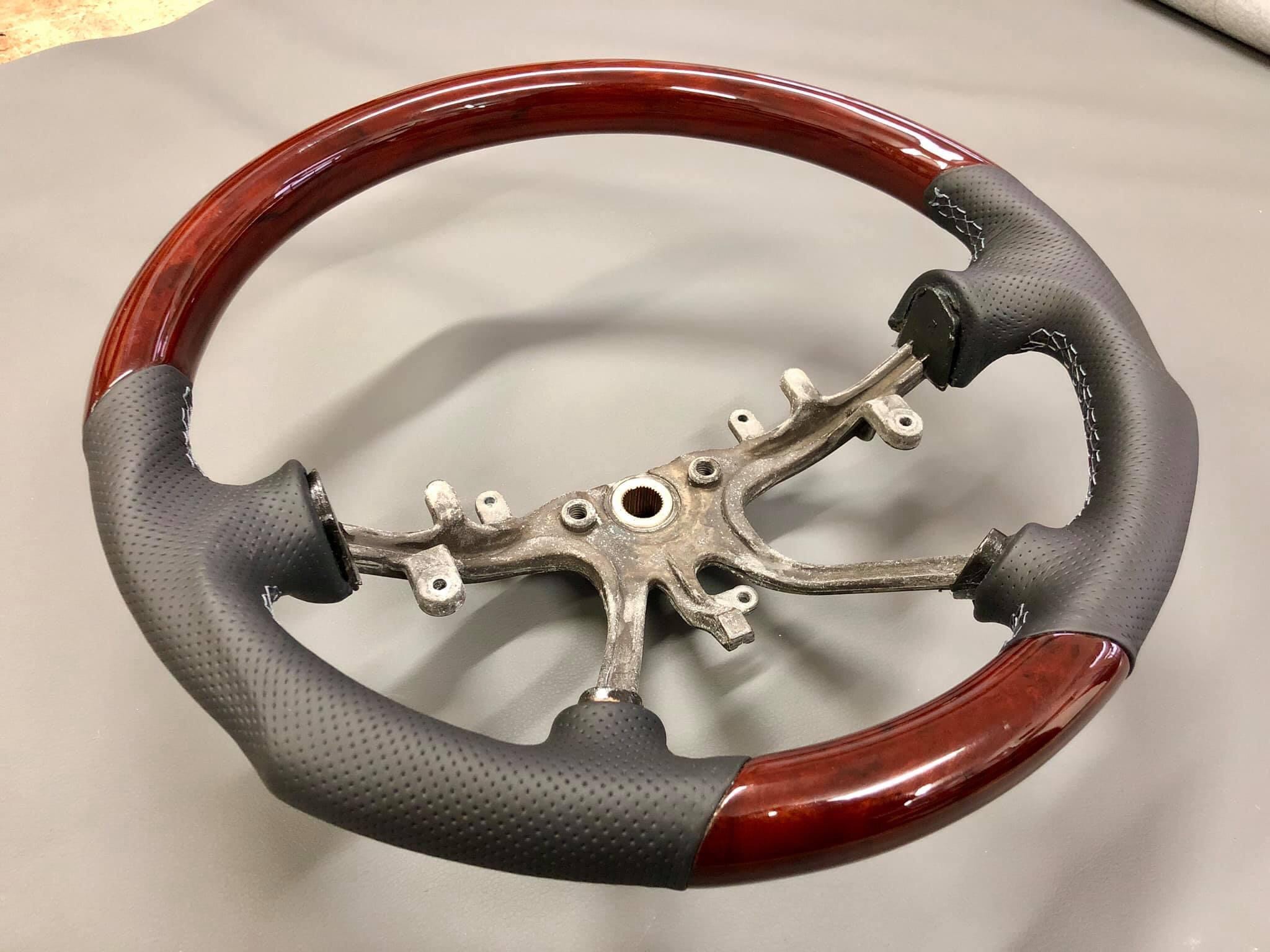 Odnowiona kierownica samochodowa: drewniany wieniec z mahoniowym połyskiem, obszyty czarną perforowaną skórą z białymi szwami, metalowy szkielet.