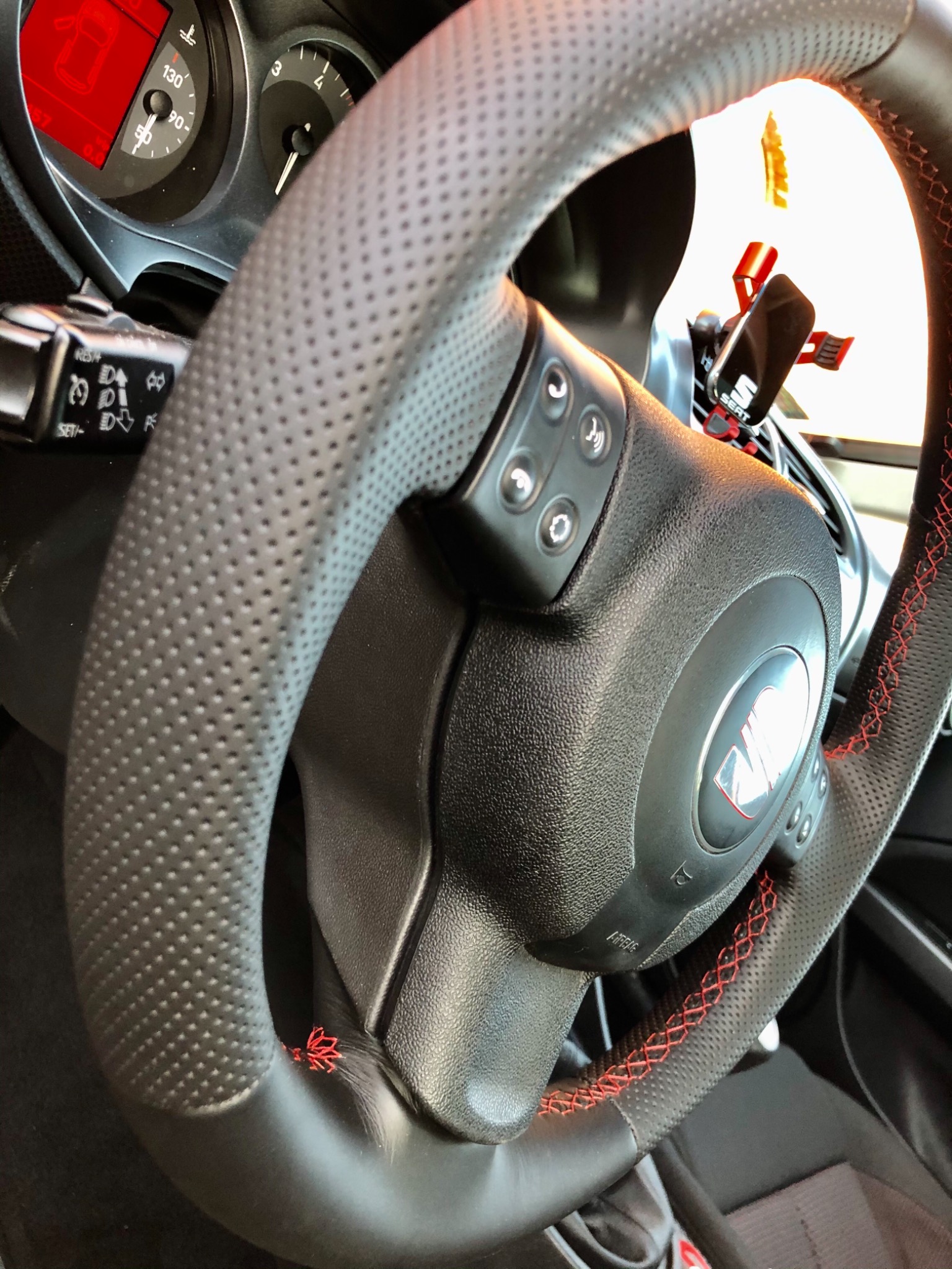 Czarna, perforowana skórzana kierownica samochodowa z czerwonym, ozdobnym szwem krzyżykowym, widoczne przyciski sterowania i logo marki.