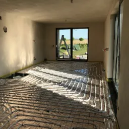 Niedokończona instalacja ogrzewania podłogowego w nowym budynku, rury ułożone na macie z widocznym wyjściem na zewnątrz przez otwarte drzwi balkonowe, drabina oparta o framugę.